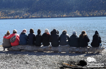 A group of people receiving mental health care therapy sitting on a log near a lake.