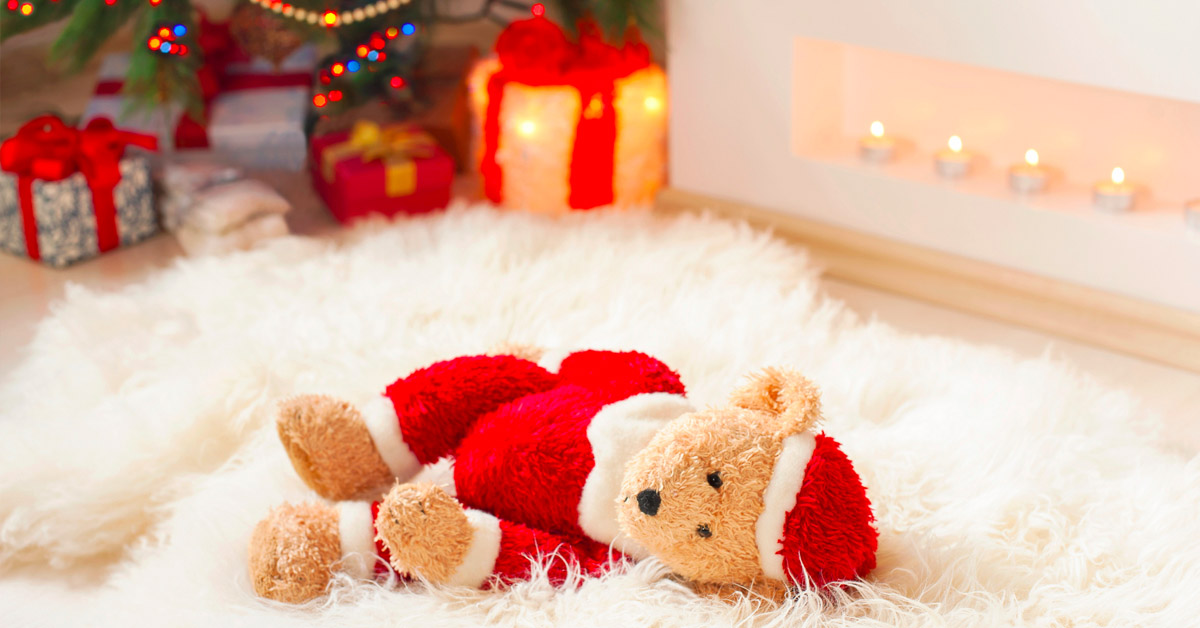 A Santa Claus teddy bear laying on a white rug in front of a Christmas tree.